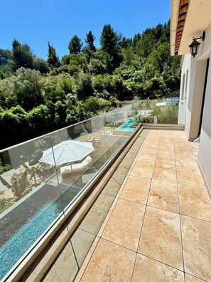 A glass balustrade overlooking a swimming pool with a tiled deck, surrounded by lush greenery.