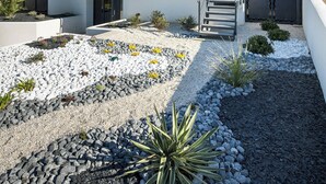 Une maison moderne à deux étages, dotée d’un balcon, d’un escalier et d’un jardin agrémenté de rochers et de plantes.