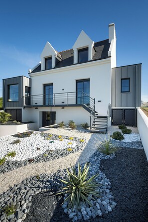 A modern two-story house with a balcony, a staircase, and a garden with rocks and plants. - Villa with spa and sea view in Finistère (Goulven)