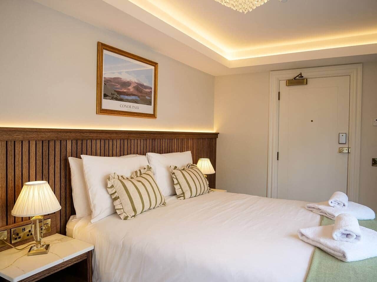 A hotel room with a bed, two bedside lamps, a framed picture on the wall, a door, and two towels on the floor.