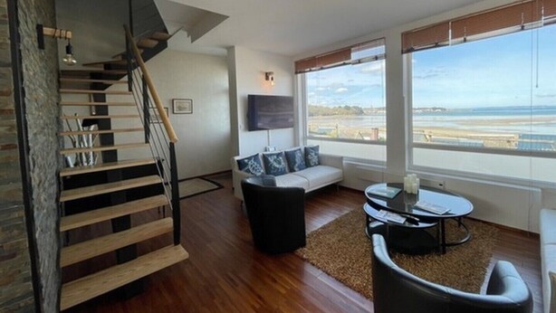 Una sala de estar moderna con una escalera, un sofá, una mesa de centro y un ventanal con vista a la playa.