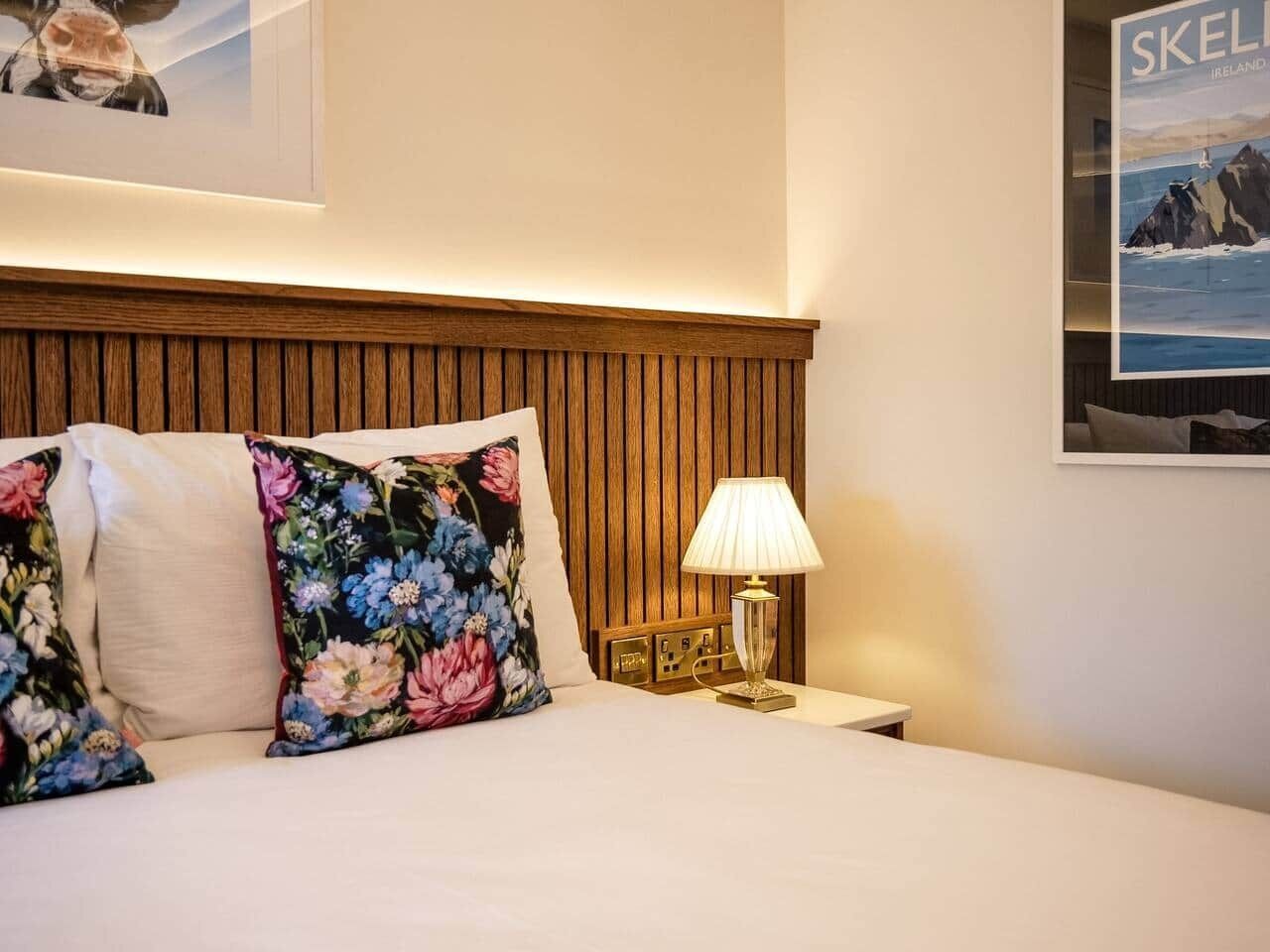 A hotel room with a wooden headboard, floral pillows, a bedside lamp, and a framed picture on the wall.