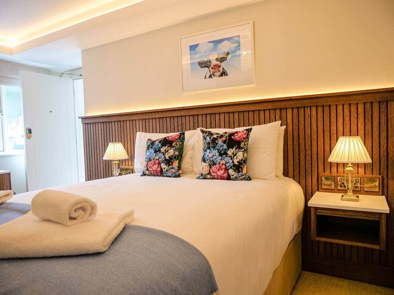 A hotel room with a bed, two pillows, a bedside table with a lamp, and a framed picture on the wall.