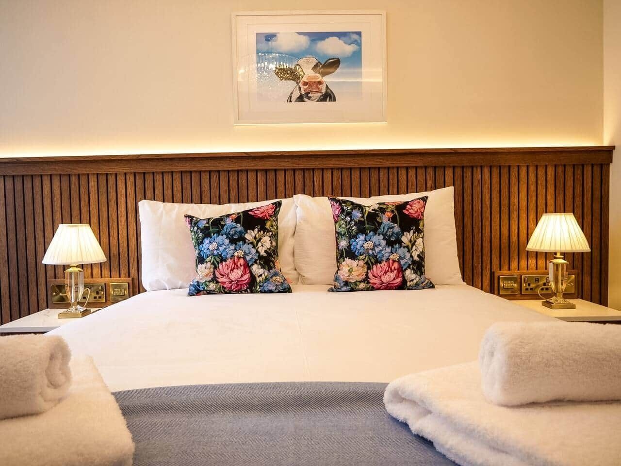 A hotel room with a bed, two pillows, two bedside lamps, a framed picture on the wall, and two towels on the bed.