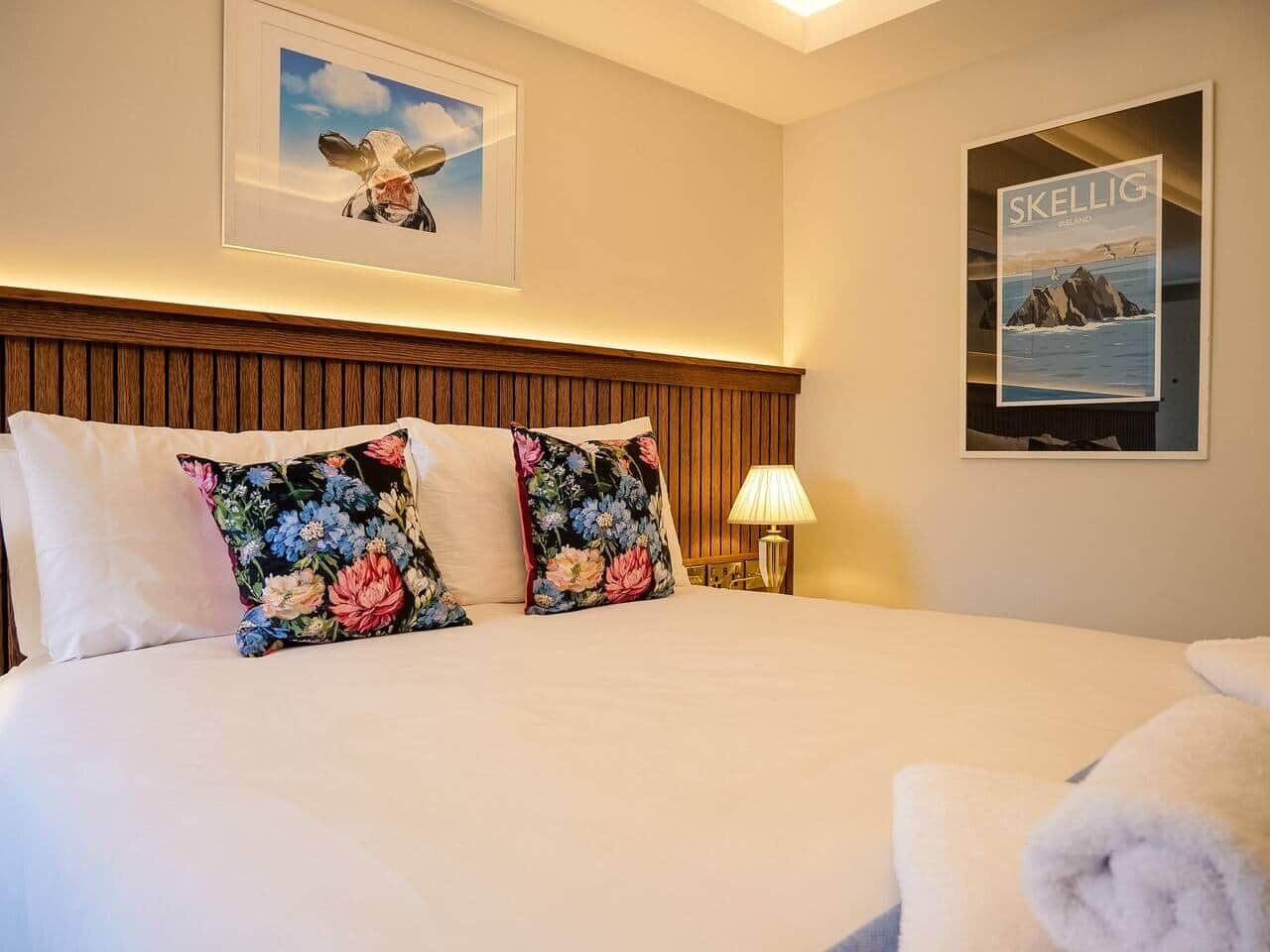 Een hotelkamer met een groot bed, bloemenkussens, een ingelijst beeld boven het bed, een lamp op het nachtkastje en een poster aan de muur.