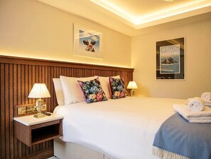 Ein Hotelzimmer mit einem hölzernen Kopfstutz, einem Bett mit weißer Bettwäsche, zwei blumigen Kissen, einem Nachttisch mit einer Lampe, einem gerahmten Bild an der Wand und einem Poster.