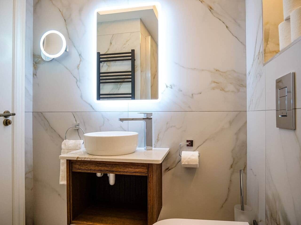 Un baño con pared de mármol, un espejo redondo con luz, tocador de madera, lavabo blanco y perchero para toallas.