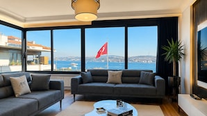 A modern living room with a sofa set, a glass coffee table, and a view of the sea with a flag. - Serene Stylish Central Cihangir Flat w/ Bosphorus 3-bedroom apartment (Beyoglu)