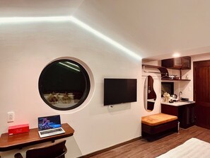 A compact room with a round window, a flat-screen TV, a laptop, a small table, and a bench. - Cocoon Danang Airport Hotel & Apartment (Da Nang)