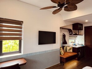 A compact living space with a flat-screen TV, a ceiling fan, a window with blinds, a bench, and a kitchenette. - Cocoon Danang Airport Hotel & Apartment (Hoa Cuong)