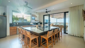 A modern kitchen with a central island, wooden chairs, ceiling fans, and a view through the glass door. - Friendly Villa (Badung)