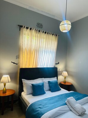 Una cama bien hecha con ropa de cama azul, dos lámparas de noche, una lámpara colgante y un reloj de pared.