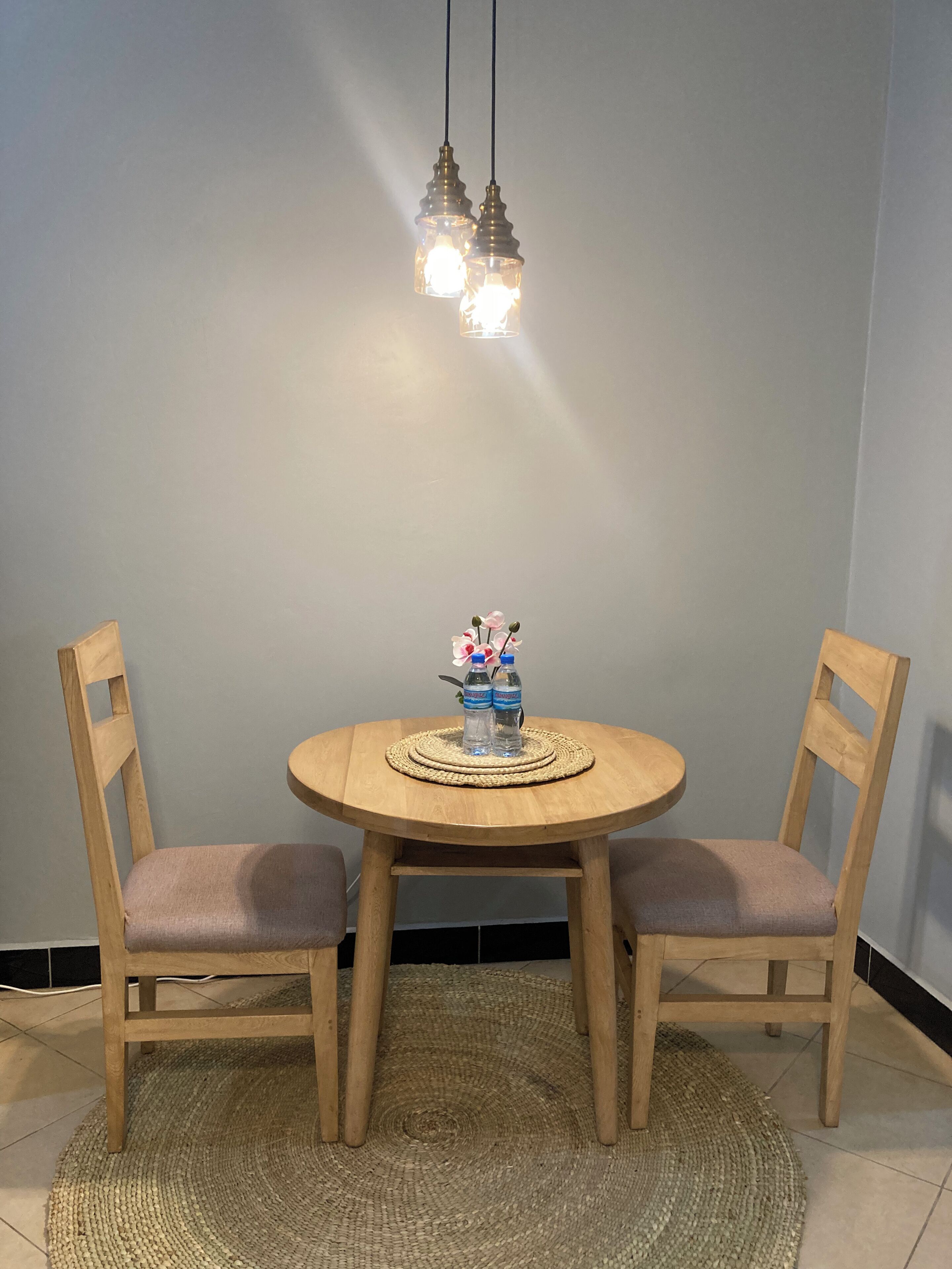 A round wooden dining table with two chairs, a woven placemat, and a vase with flowers and water bottles.