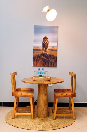 Una mesa redonda de madera con dos sillas de madera, un mantel tejido, un cuadro enmarcado con un león, y dos botellas de agua.
