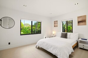 Ein Schlafzimmer mit einem großen Bett, zwei Nachttischen, einem Beistelltisch, einem runden Spiegel und zwei Fenstern mit Blick auf Grünflächen.