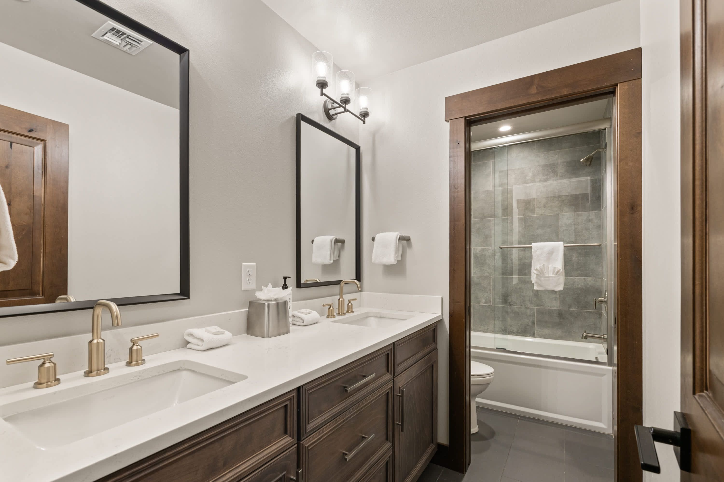 A modern bathroom with a double sink, a large mirror, a shower, and a wooden door.