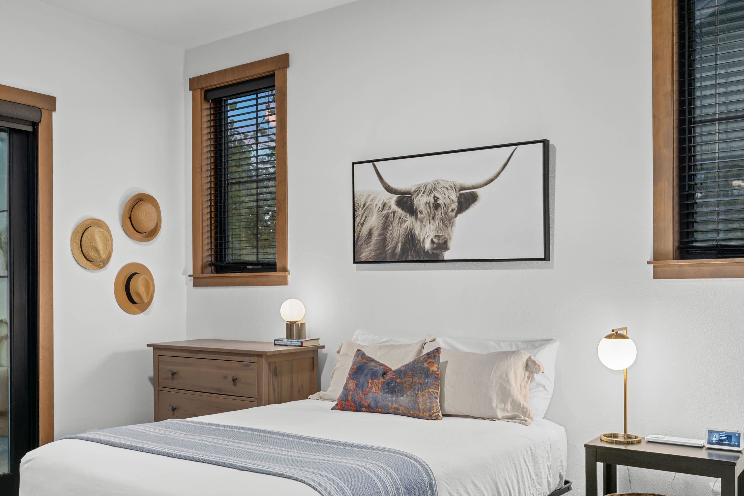 A bedroom with a bed, a nightstand, a lamp, a framed picture of a bull, and three hats on the wall.