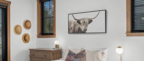 A bedroom with a bed, a nightstand, a lamp, a framed picture of a bull, and three hats on the wall.