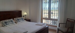 Hotelkamer met een bed, nachtkastje met lamp, raam met uitzicht op palmbomen en een balkon, plus een houten stoel.