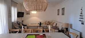 Un coin repas avec une table dressée pour deux personnes, un lustre en osier, un canapé avec des coussins, un téléviseur à écran plat et des décorations murales.
