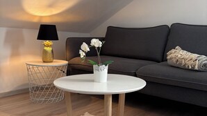 Ett mysigt vardagsrum med ett runt, vitt bord, en mörk soffa, ett litet vitt sidobord med en lampa och en växt.