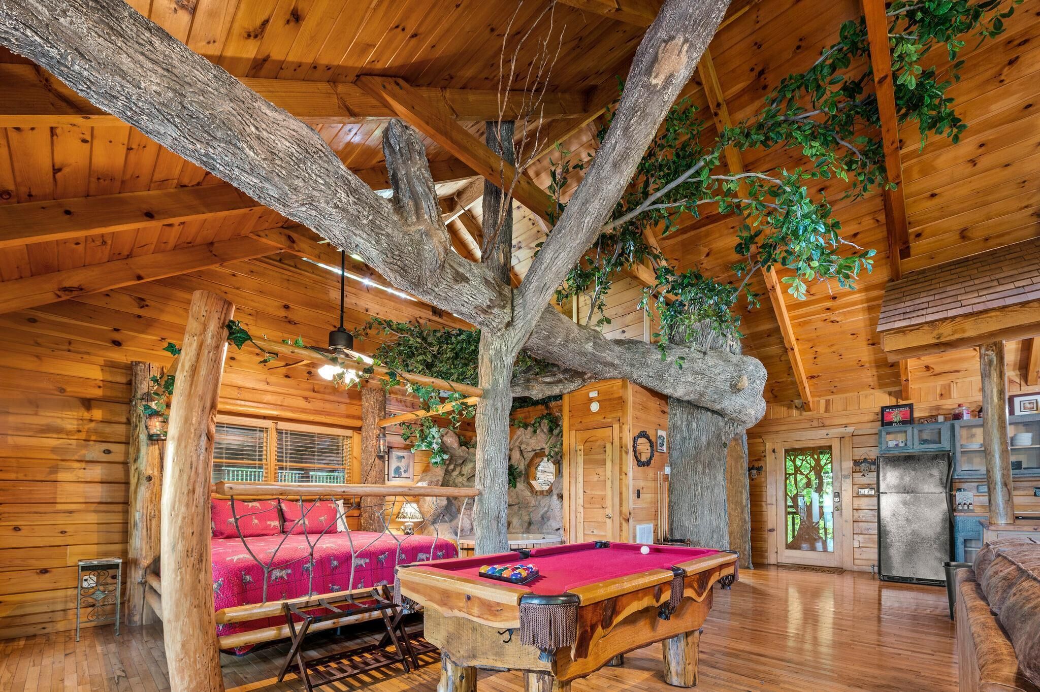 A wooden cabin interior with a pool table, a red sofa, and a large tree trunk integrated into the ceiling.