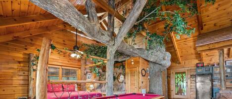 A wooden cabin interior with a pool table, a red sofa, and a large tree trunk integrated into the ceiling.