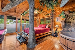 Interior de una cabaña de madera con cama, bañera de hidromasaje y vistas al exterior.
