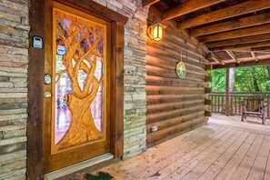 Una cabaña de madera con pared de piedra, una puerta de madera con tallado de árbol, un porche con banco y un cartel que indica zona para perros.