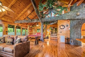 Interior de una cabaña de troncos con techo de madera, un sofá con cojines de temática animal, una mesa de billar y un gran tronco de árbol como elemento central.