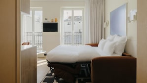 A hotel room with a bed, a TV, a balcony, and a view of buildings. - Edgar Suites - Jules Ferry (Paris)