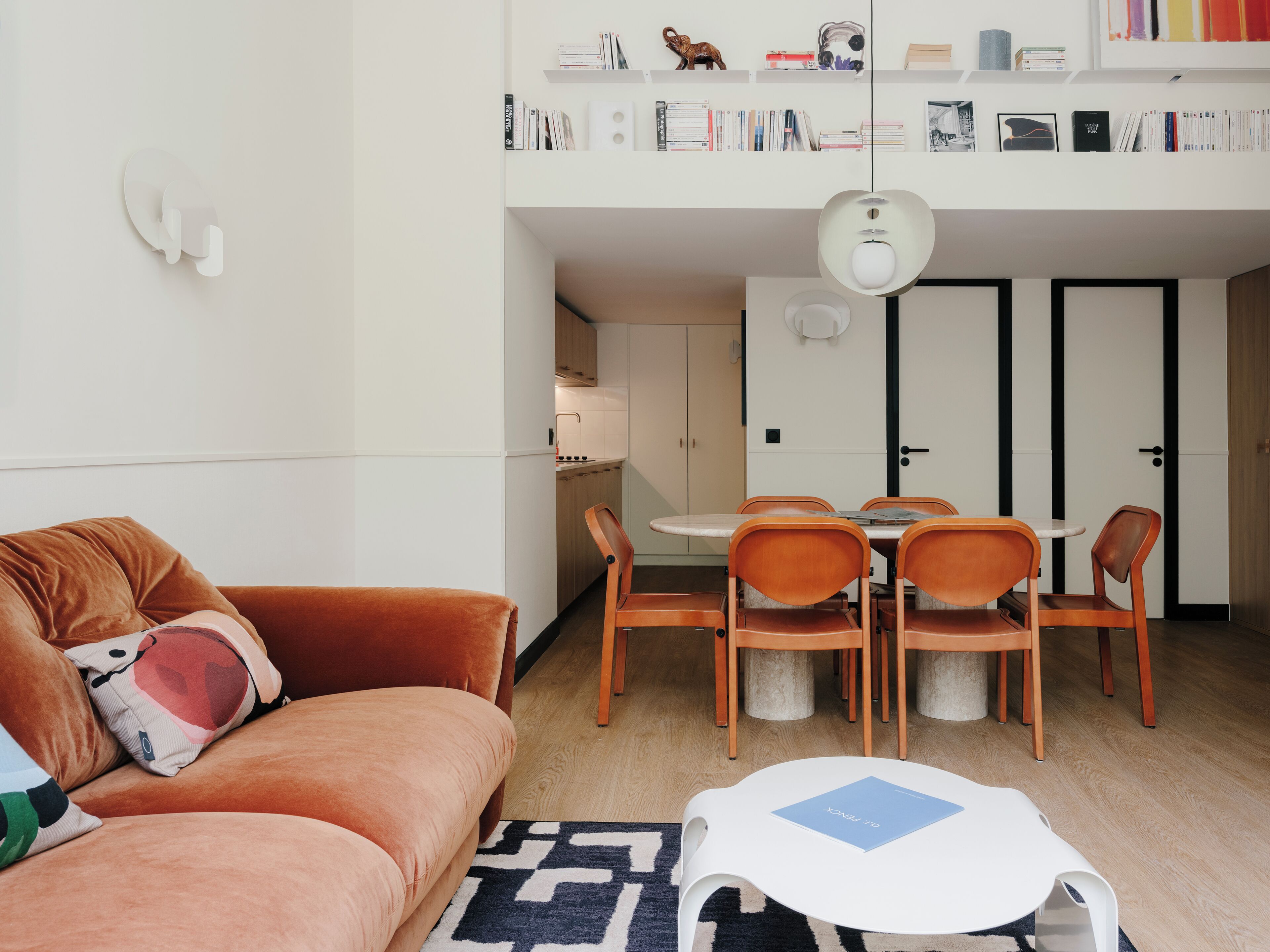 Ett kompakt bostadsutrymme med en soffa, ett matbord, stolar, ett litet kök och en bokhylla.