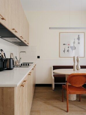 A modern kitchen with wooden cabinets, a white countertop, and a dining area with a round table and orange chairs. - Edgar Suites - Jules Ferry (Paris)
