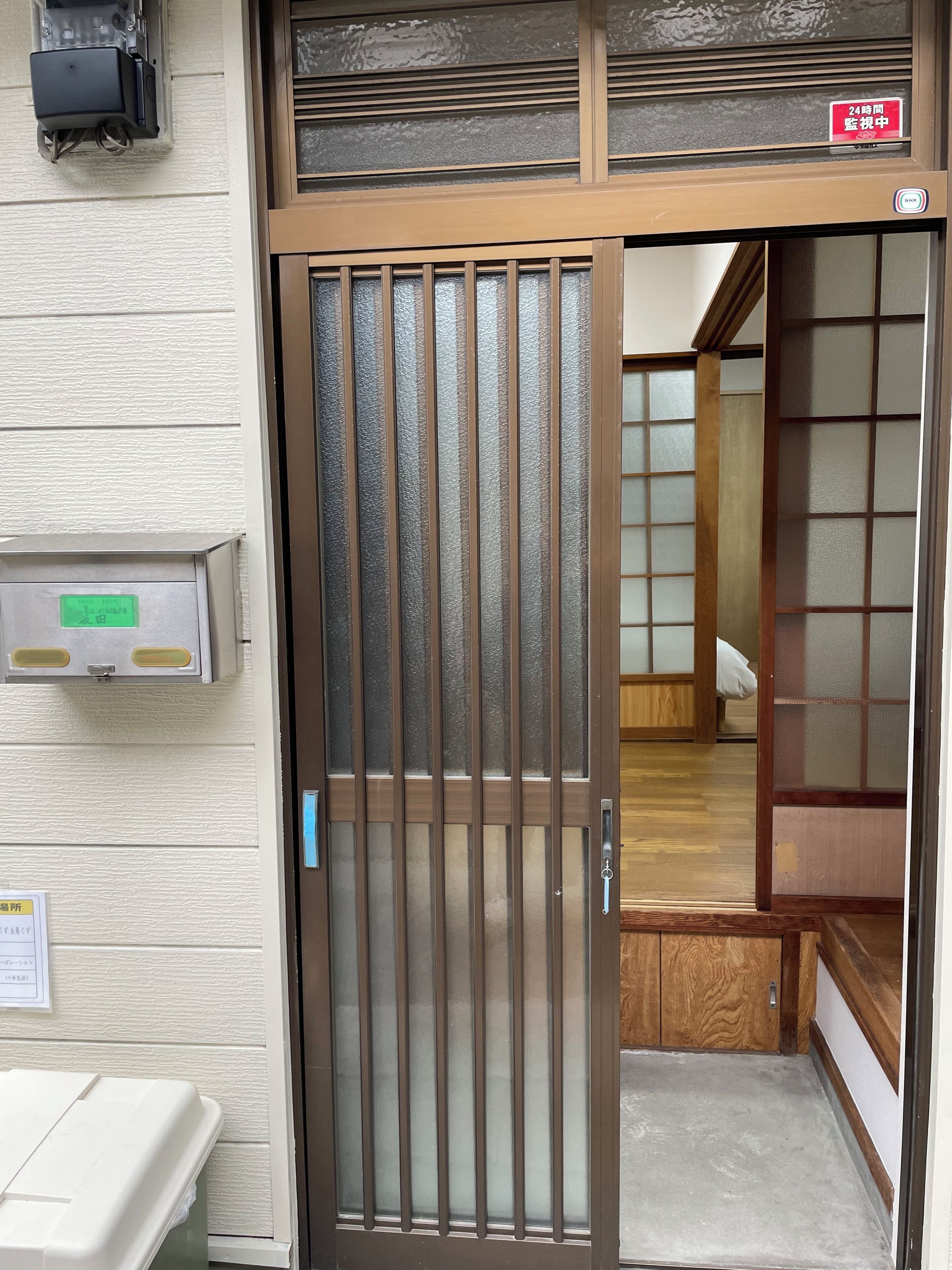 A traditional Japanese-style entrance with a sliding glass door, a wooden interior visible through the door, and a sign indicating a 24-hour safe deposit box.. House in Sumiyoshi Ward, at Kobayashi Guest House, Osaka