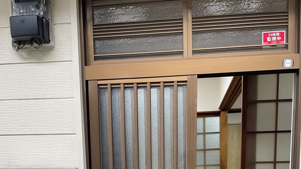 Uma entrada tradicional no estilo japonês, com porta de vidro deslizante, interior de madeira visível através da porta, e placa indicando cofre disponível 24 horas.