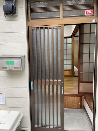 A traditional Japanese-style entrance with a sliding glass door, a wooden interior visible through the door, and a sign indicating a 24-hour safe deposit box.. House in Sumiyoshi Ward, at Kobayashi Guest House, Osaka