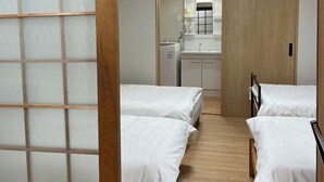 Quarto com duas camas, piso de madeira, pequeno espaço de cozinha e porta deslizante com painéis de vidro fosco.