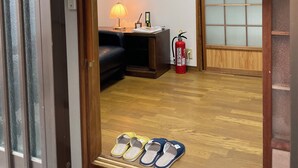 Um quarto com piso de madeira, um sofá preto, uma luminária, um extintor de incêndio e chinelos em uma prateleira.