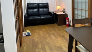 Quarto com sofá de couro preto, mesa de jantar de madeira, pequena mesa lateral com abajur e extintor de incêndio.