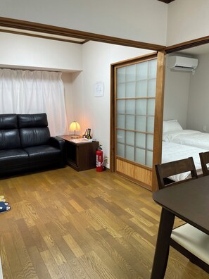 Une piÚce avec un plancher en bois, un canapé en cuir noir, une petite table avec une lampe, un extincteur et un lit avec un cadre en bois.