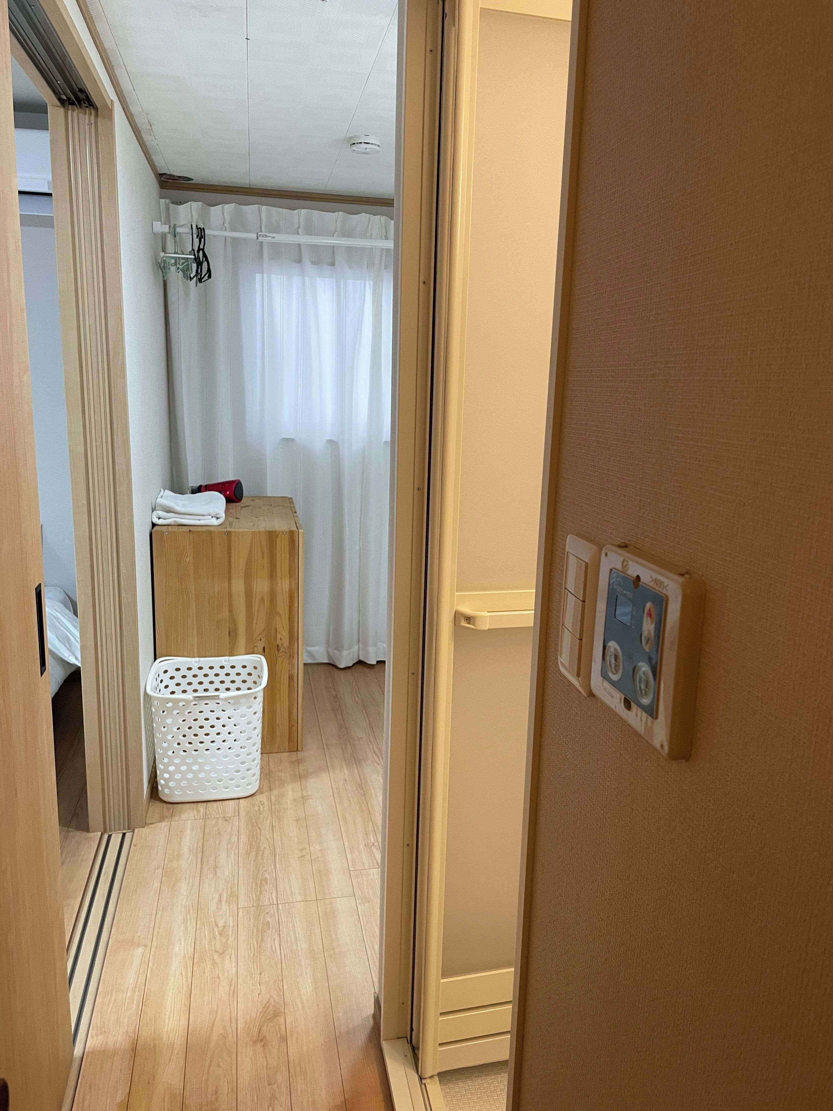 A room with a wooden floor, a white laundry basket, a small table with a red object, and a white curtain.