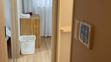 A room with a wooden floor, a white laundry basket, a small table with a red object, and a white curtain.
