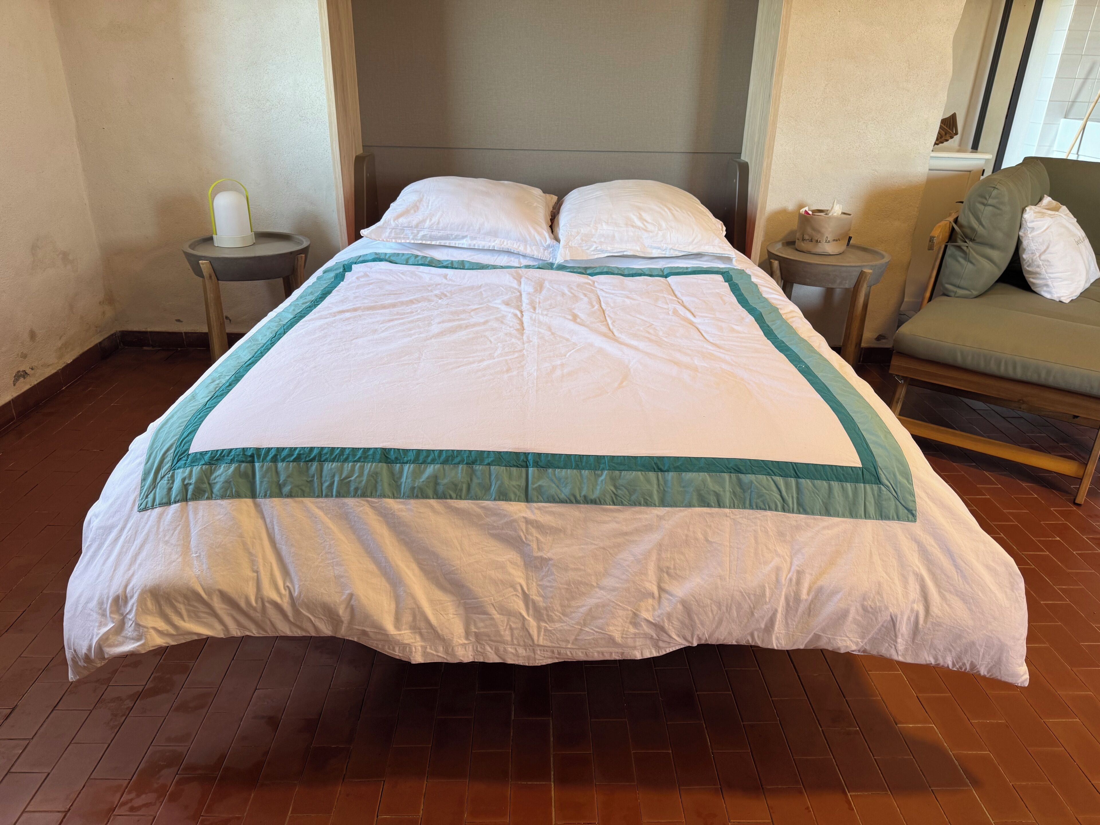 Un lit bien fait, recouvert d’une couette blanche et turquoise, une table de chevet avec une lampe, et une chaise agrémentée de coussins.