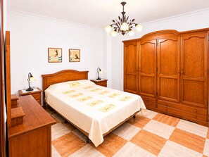 A bedroom with a bed, nightstands, a wooden wardrobe, a chandelier, and two wall-mounted pictures. - Apartamento San Jose bajo (Conil de la Frontera)