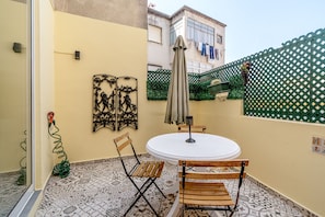 Pequeno alpendre exterior com uma mesa redonda e duas cadeiras de madeira, uma escultura de metal e uma vedação verde.