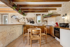 A rustic kitchen with wooden beams, a stone wall, and a dining area with a wooden table and chairs.