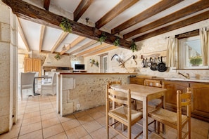 A rustic kitchen with exposed wooden beams, a stone wall, and a dining area with wooden chairs and a table.