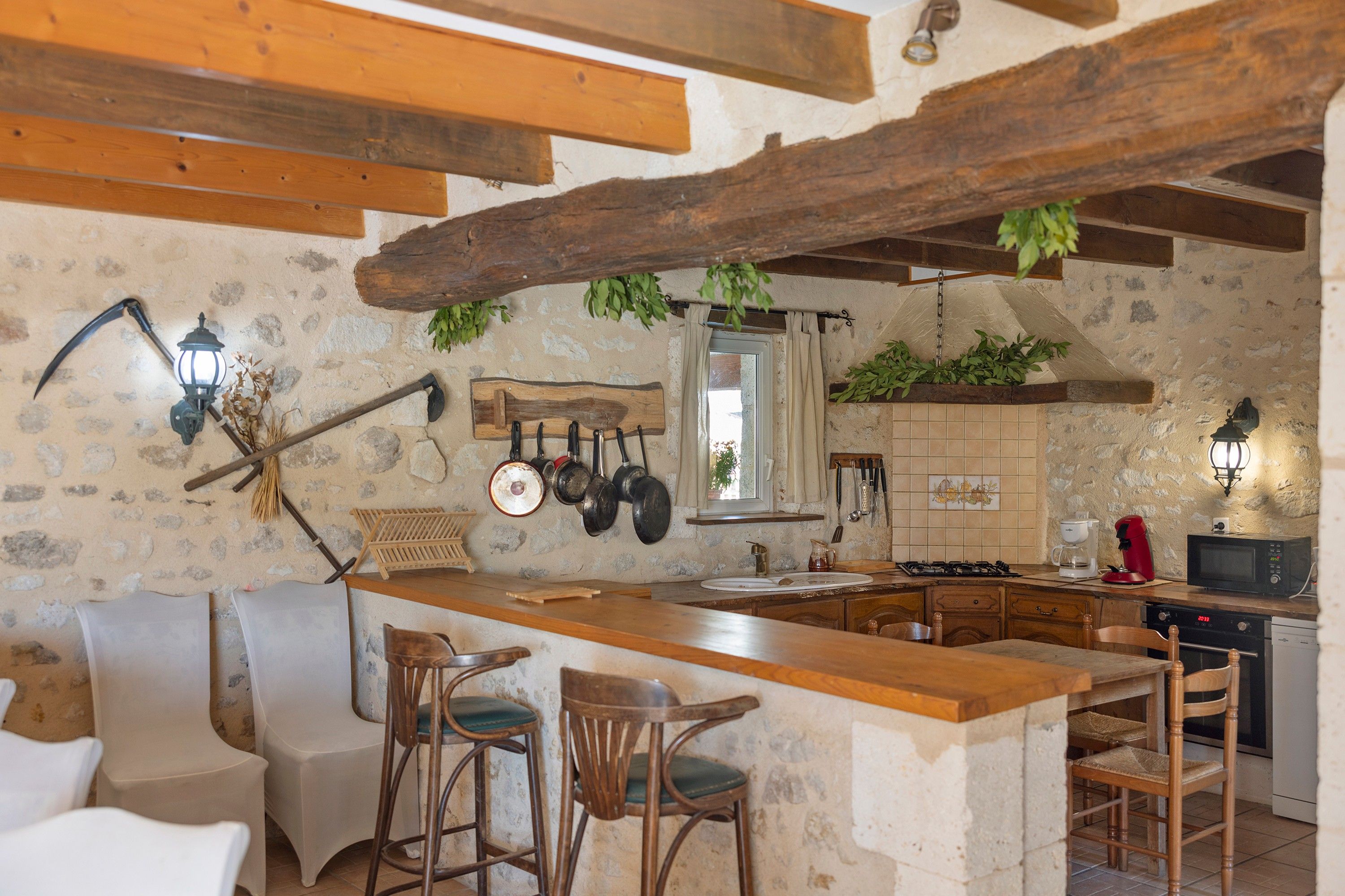 Una cocina rústica con paredes de piedra, vigas de madera, una zona de bar con taburetes y varios utensilios de cocina colgados en la pared.