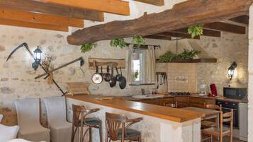 Una cocina rústica con paredes de piedra, vigas de madera, una zona de bar con taburetes y varios utensilios de cocina colgados en la pared.