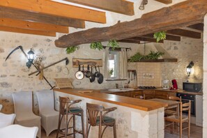 A rustic kitchen with stone walls, wooden beams, a bar area with stools, and various kitchen utensils hanging on the wall. - Country house 'Seul Au Monde' with private pool, terrace and private garden (Guizengeard)